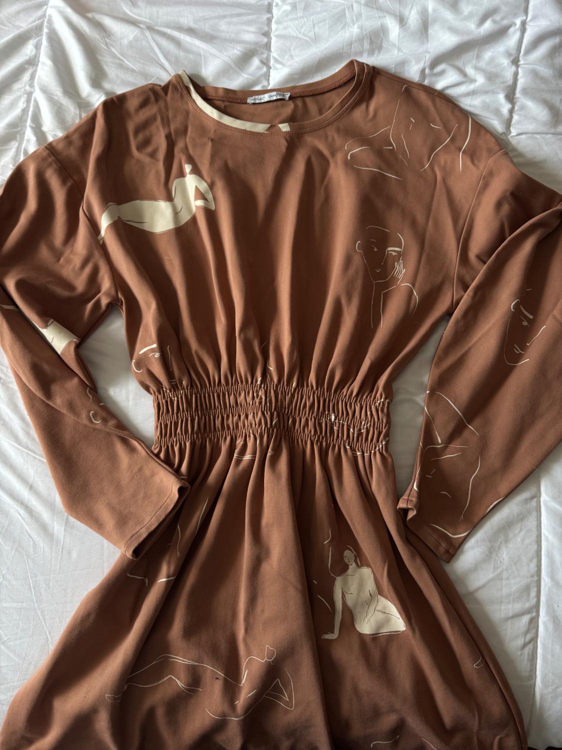 Brown dress with abstract white human figure print, long sleeves and ruched waist, on a white bedspread.