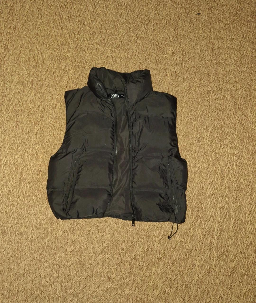 Black padded gilet on a beige carpet, with a front zip fastening and high neck. Visible label inside the collar.