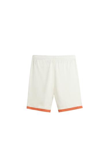 Back view of white Bermuda shorts with elastic waistband and orange bands on the bottom edge.