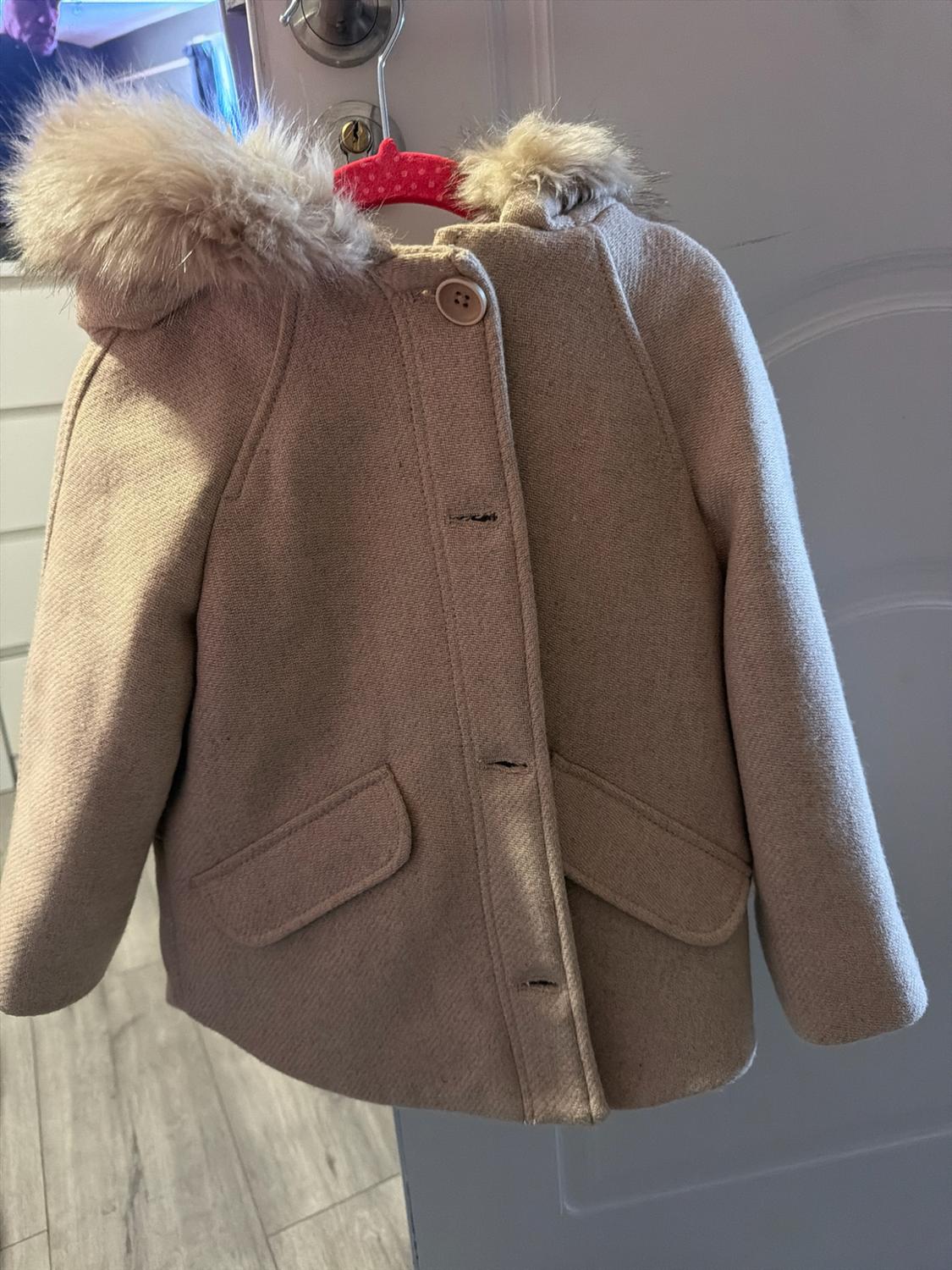 Beige coat with fur-lined hood and front pockets, hanging on a pink clothes hanger from a white door handle.