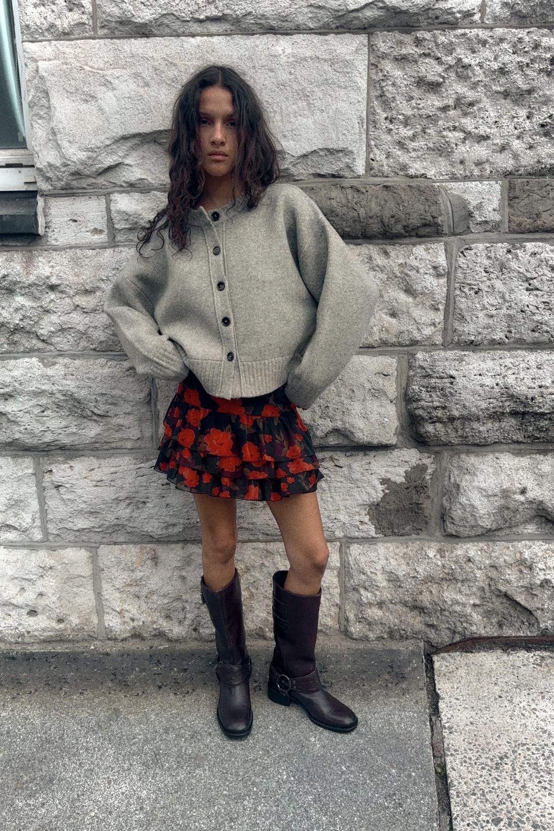 Siv komplet s pleteno jopico na gumbe, kratko krilo z volančki in rdečim cvetličnim potiskom, visoki rjavi škornji.