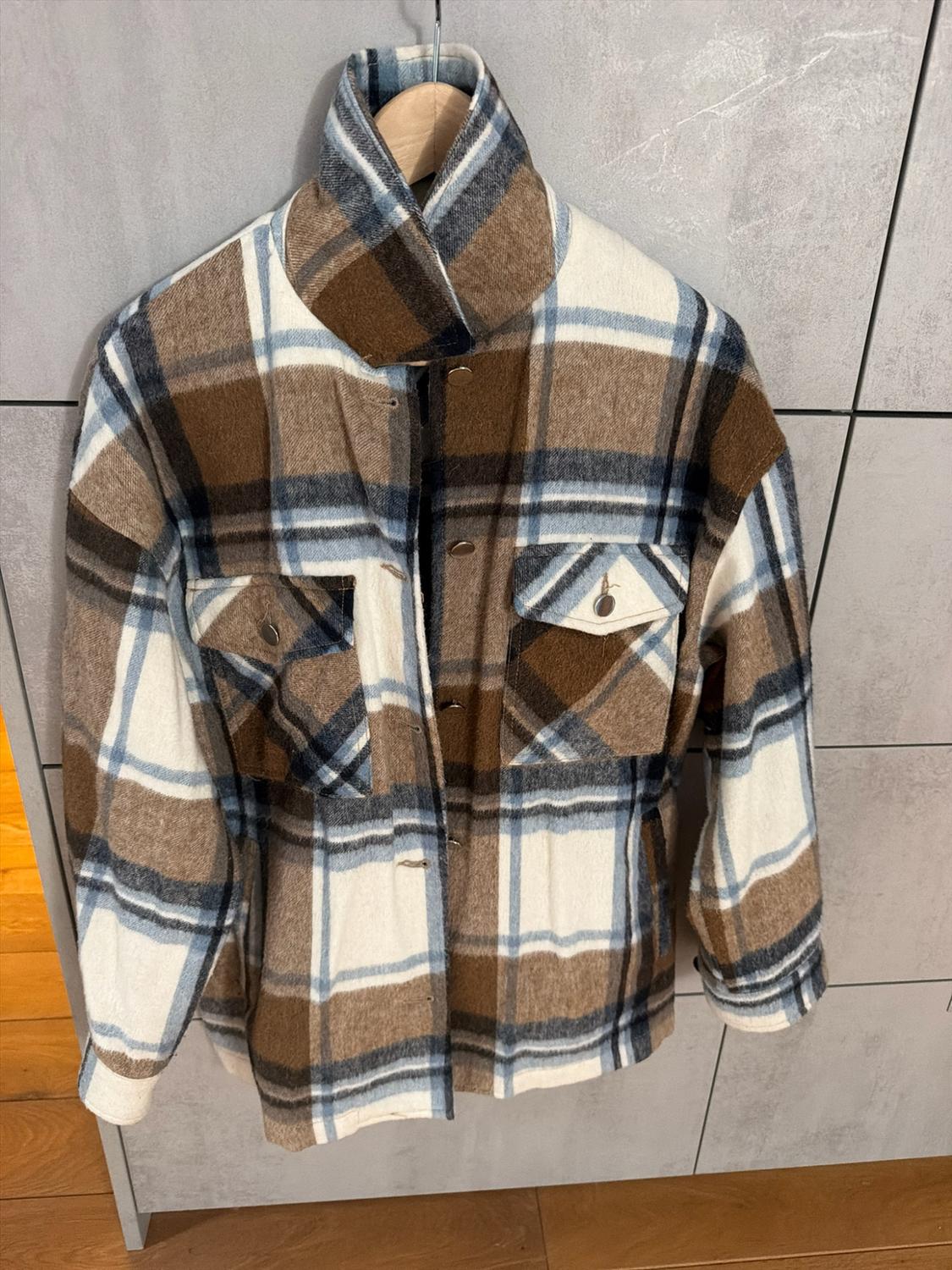 Brown, white and blue check overshirt with pockets, hanging on a wooden hanger against a grey wall.