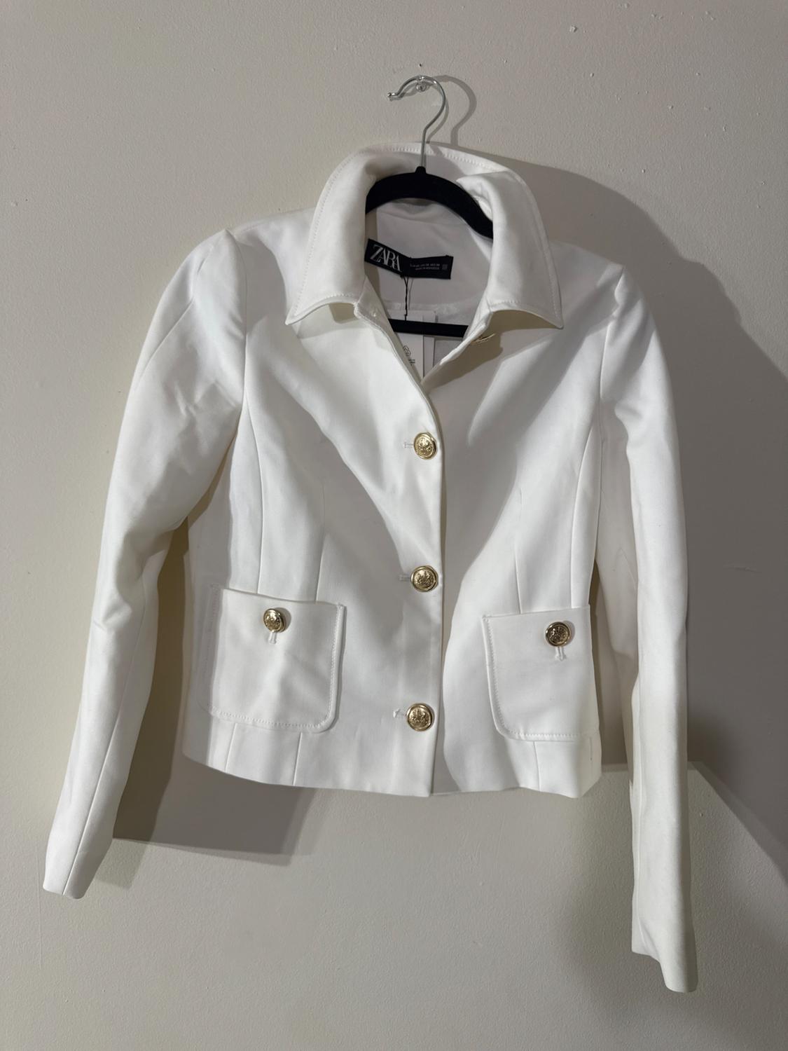 White women's jacket with gold buttons and front pockets, hanging on a black hanger against a light wall.