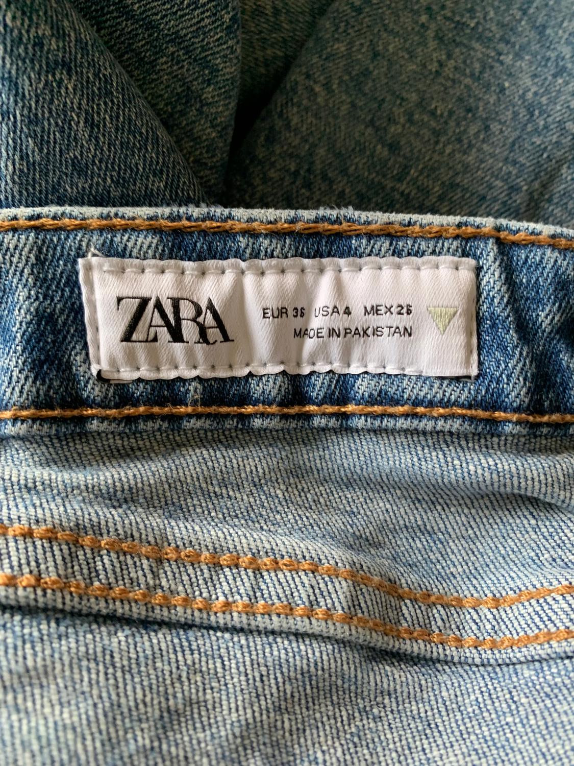 Jeans label with text: "ZARA, EUR 36, USA 4, MEX 26, MADE IN PAKISTAN". Blue denim fabric with visible orange stitching.