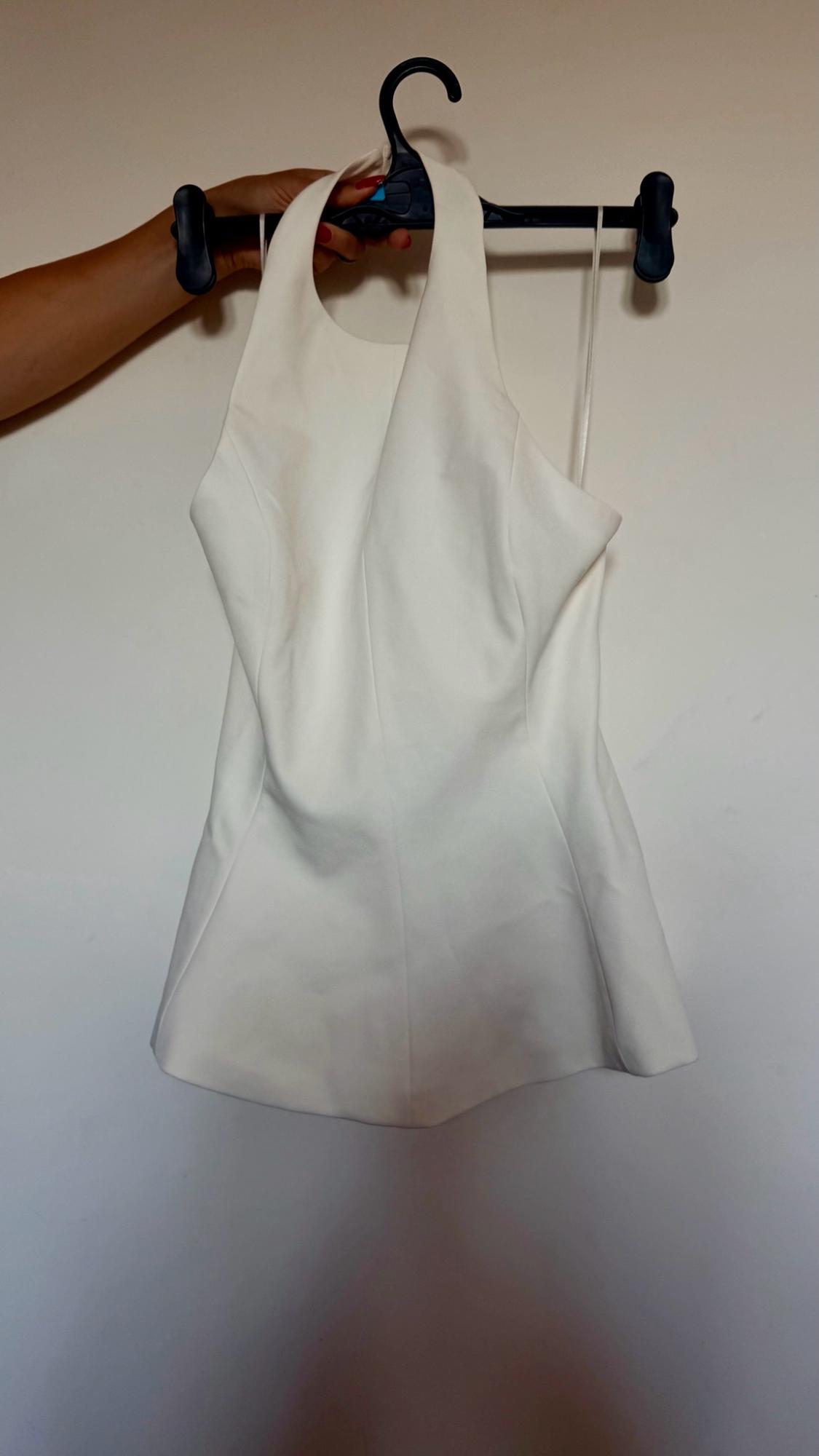 Hand with red nails holding a white sleeveless top on a black hanger against a light wall. Casual style.