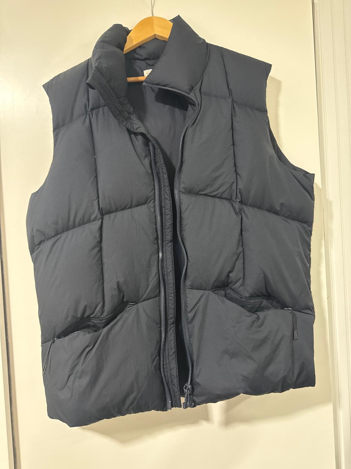Black padded gilet hanging on a wooden hanger, with a front zip and side pockets with zips.