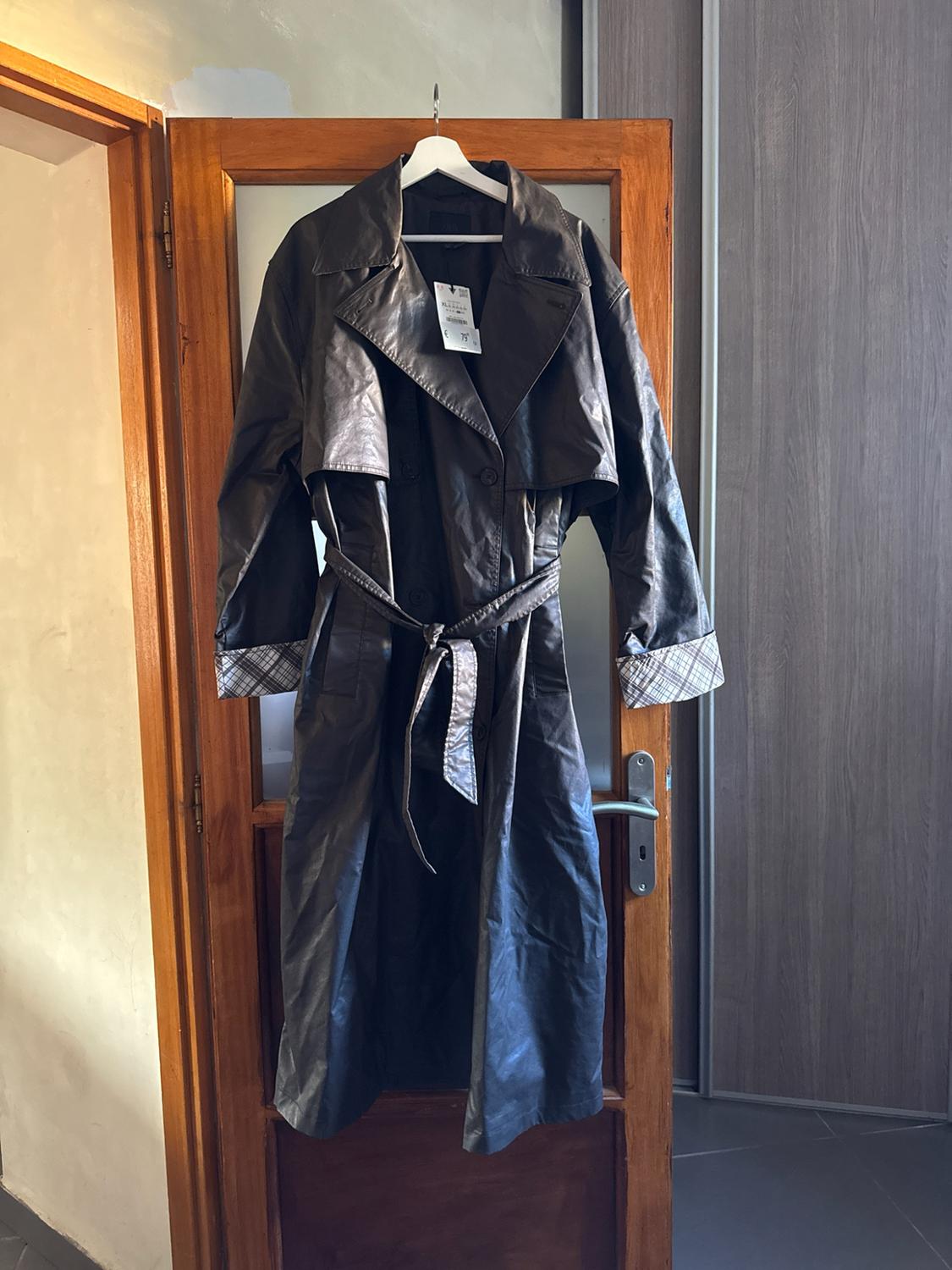 Long brown trench coat with a belted waist and checked cuffs, hanging on a hanger over a glazed wooden door.