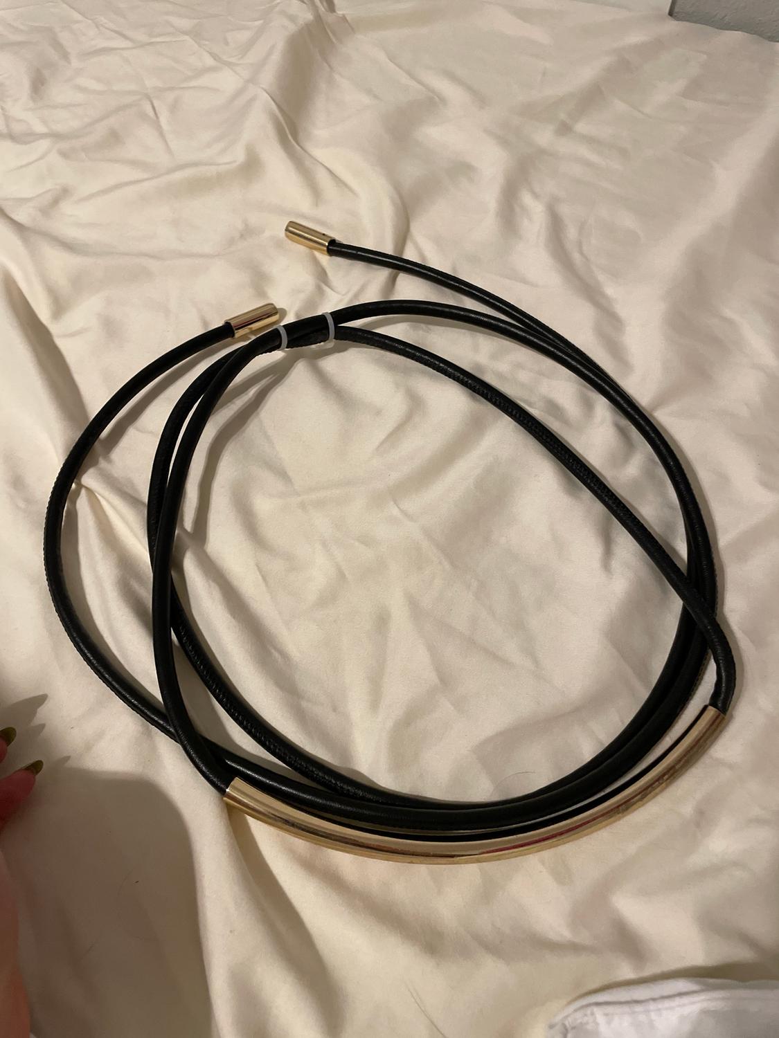 A black belt with gold details lies on a crumpled white fabric. In the bottom left corner, a hand with long, painted nails appears.