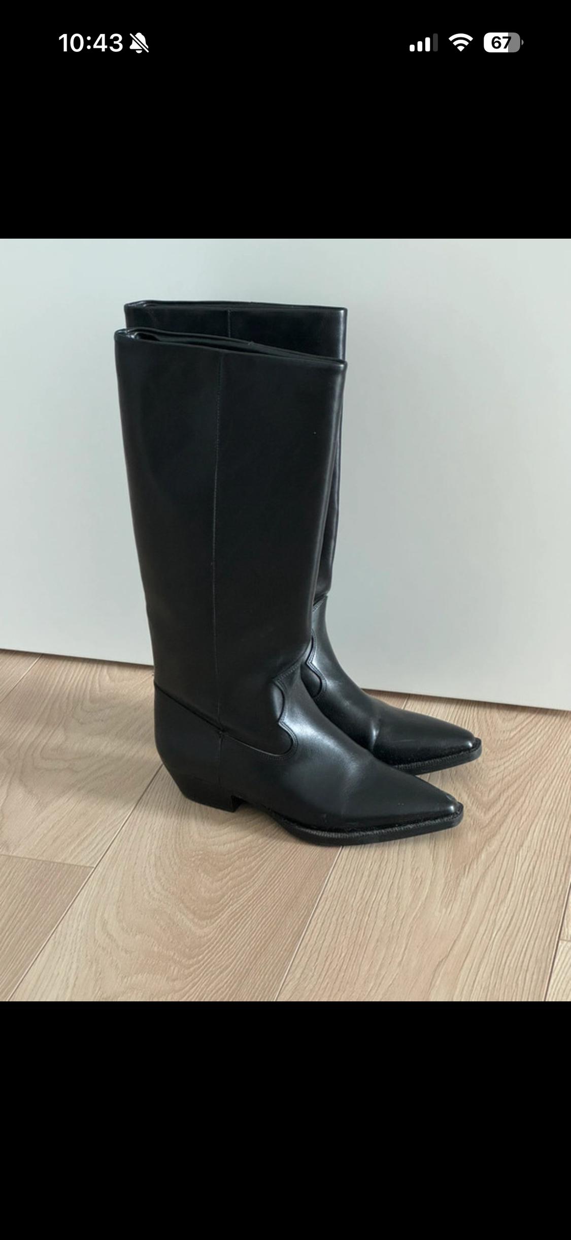Black knee-high boots with a pointed toe on a light wooden floor in front of a white wall. Text: 10:43 67.