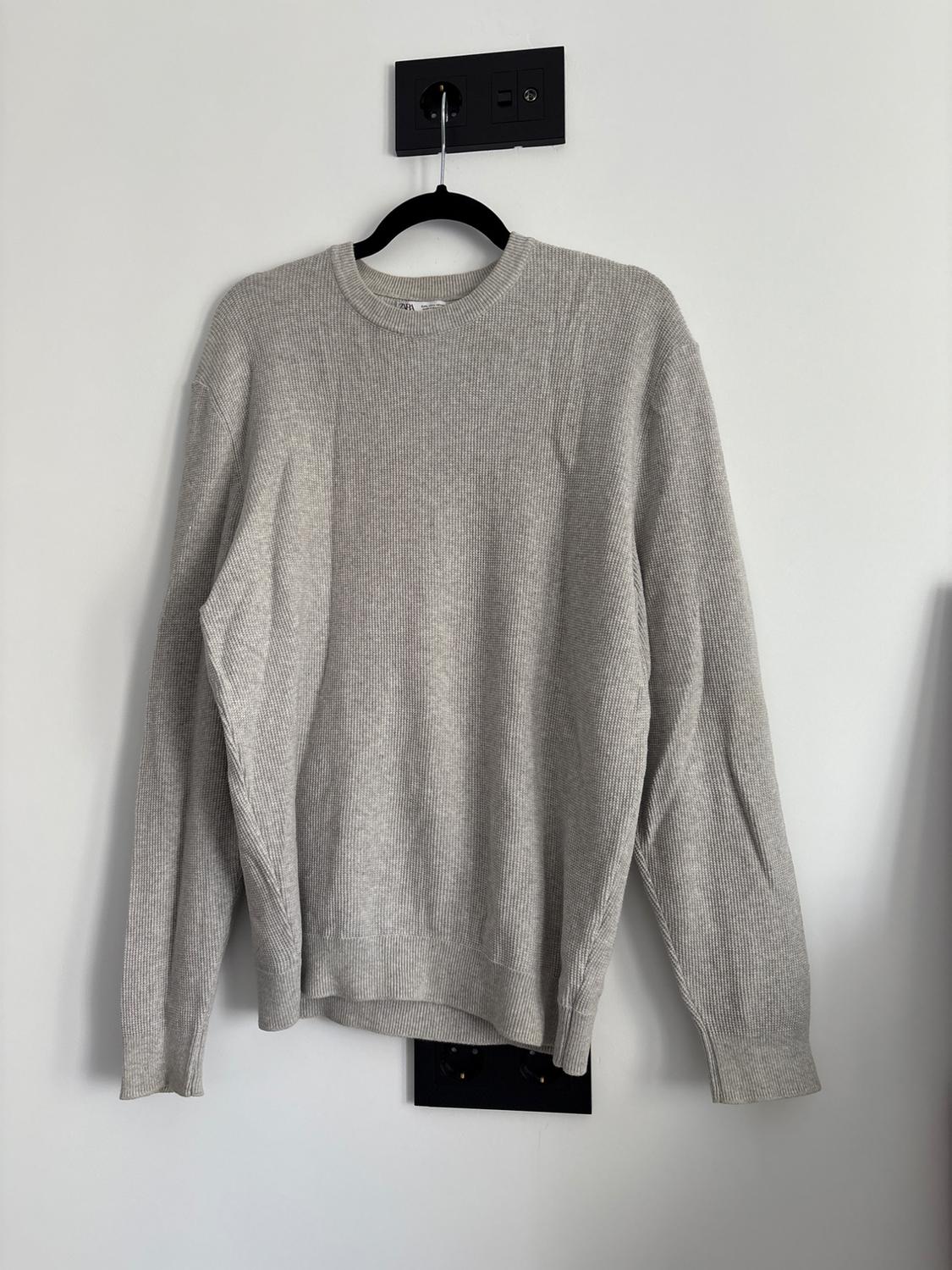 Light grey ribbed jumper with a crew neck and long sleeves, hanging on a black hanger against a white wall with black sockets.
