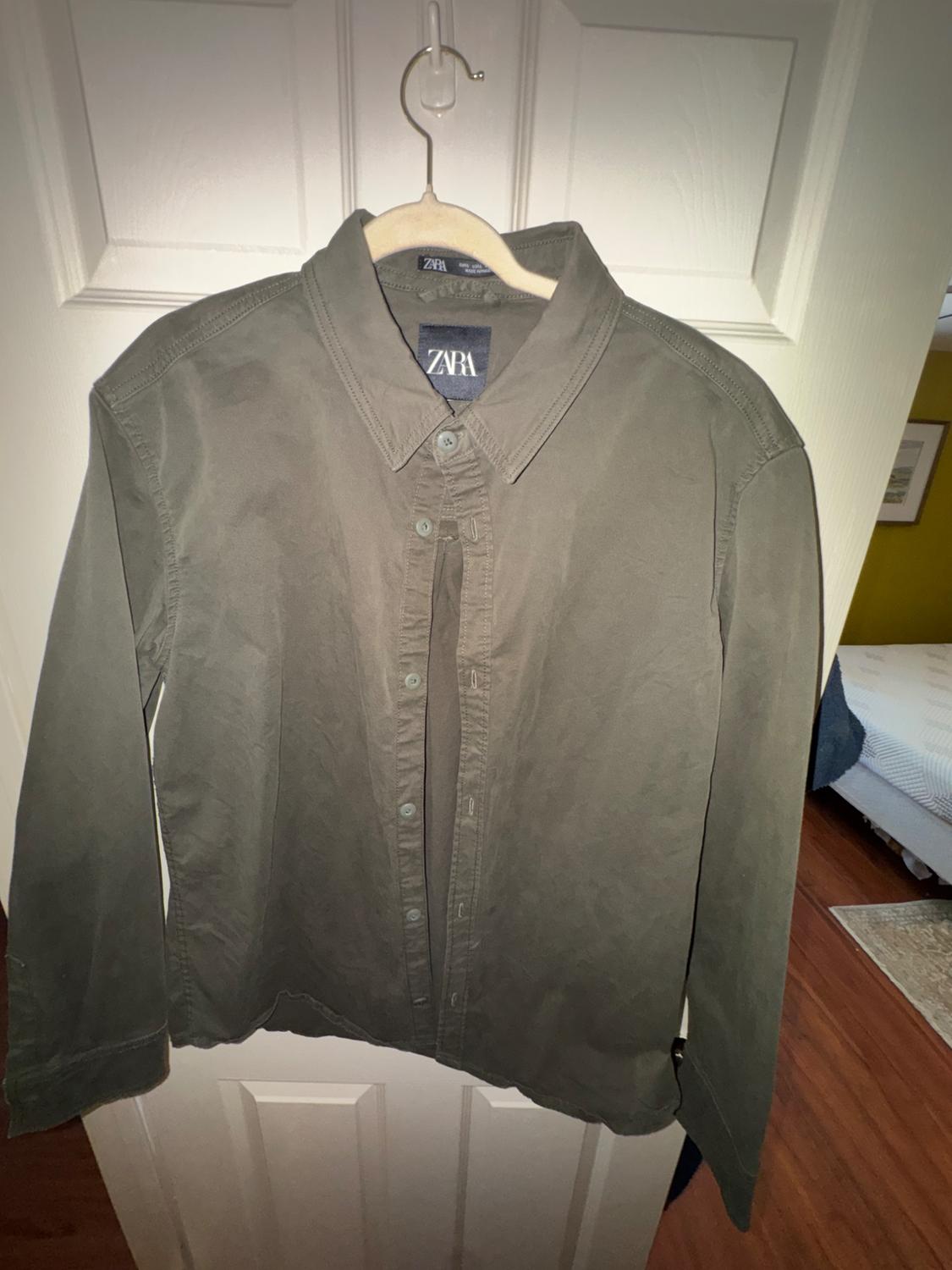 Olive green long-sleeved shirt hanging on a wooden hanger, with a visible Zara tag, over a white door.