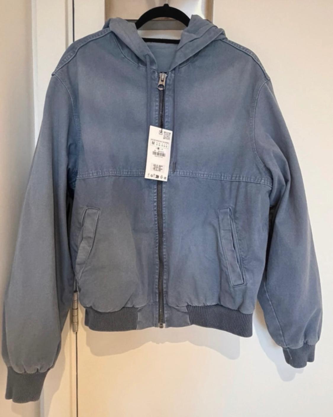 Faded blue hooded jacket with a zip and label, hanging on a black hanger against a white door.