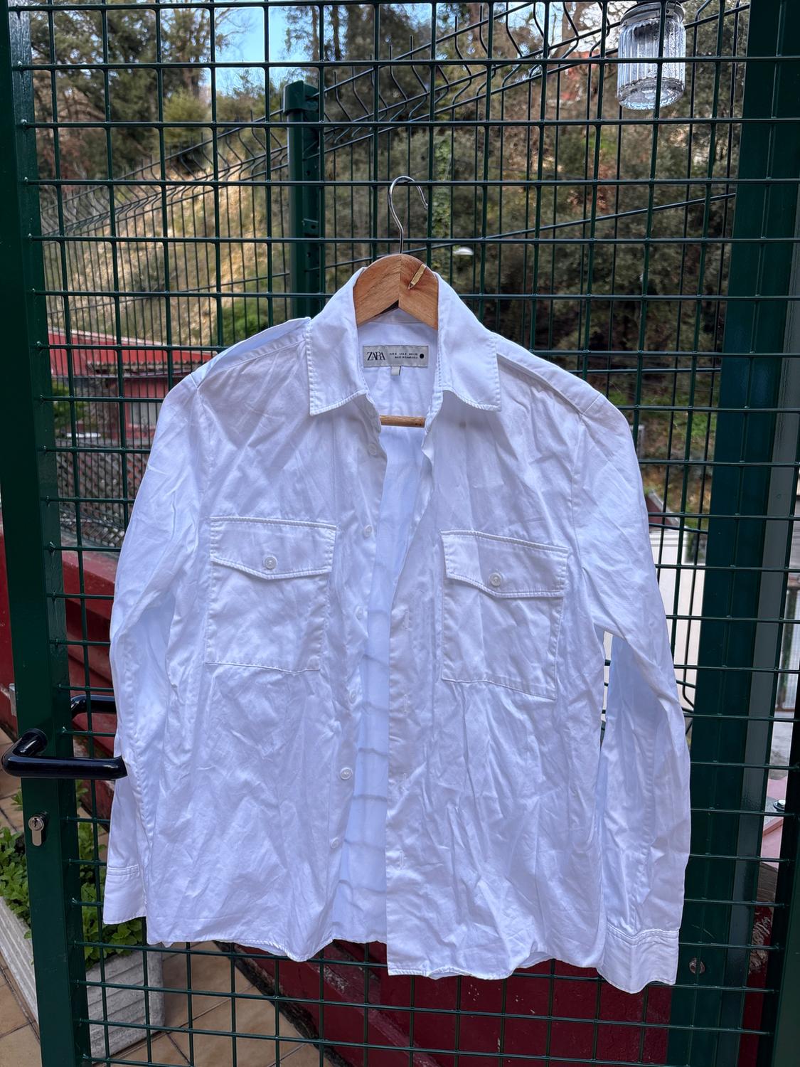 Wrinkled white shirt hanging on a wooden hanger over a green fence. Visible label with "ZARA" text. Garden background.