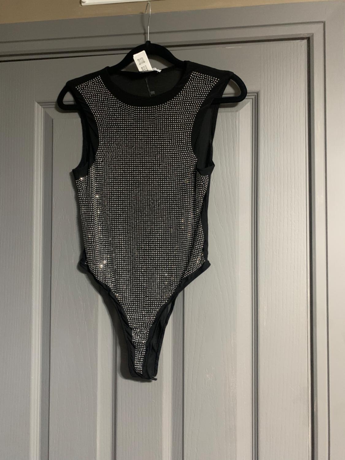 Sleeveless black bodysuit with sparkly details, hanging on a grey door. Label visible on the hanger.