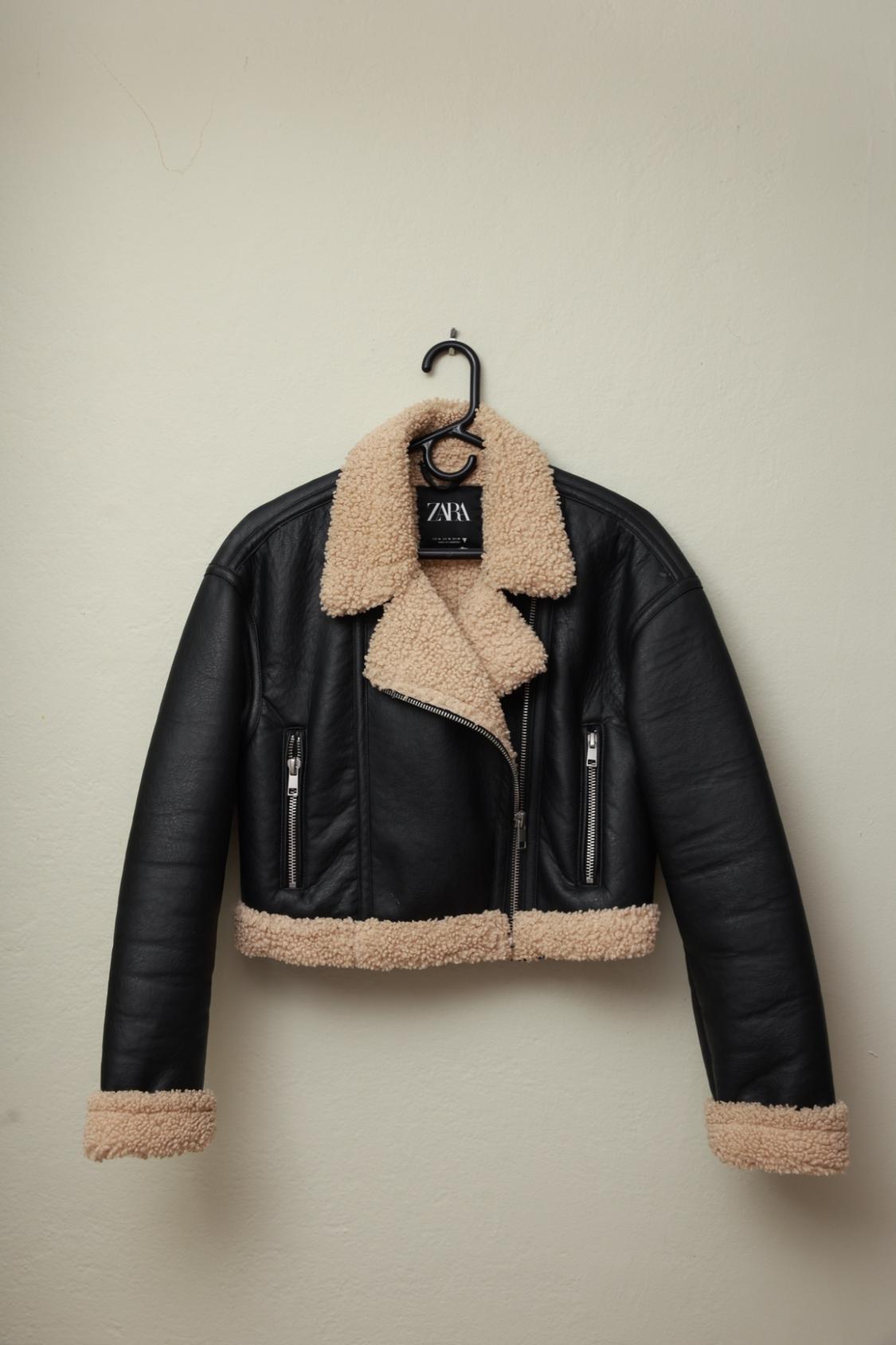 Black jacket with beige faux fur collar, hem, and cuffs, hanging on a hanger against a light-colored wall. Text: ZARA.