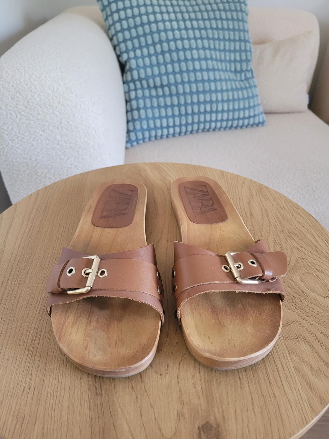 Brown sandals with a wooden sole and buckle on a wooden table. ZARA text. Relaxed atmosphere, background with a white sofa and blue cushion.