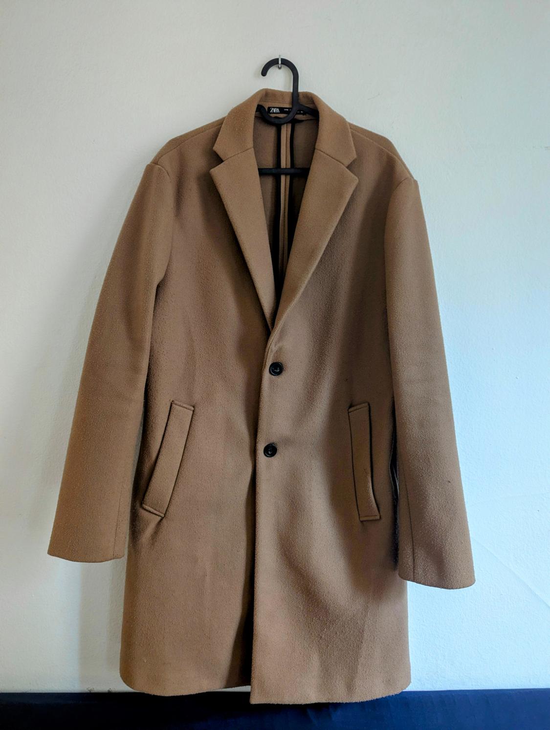 Light brown coat hanging on a black hanger against a white wall. It has lapels and two front buttons.