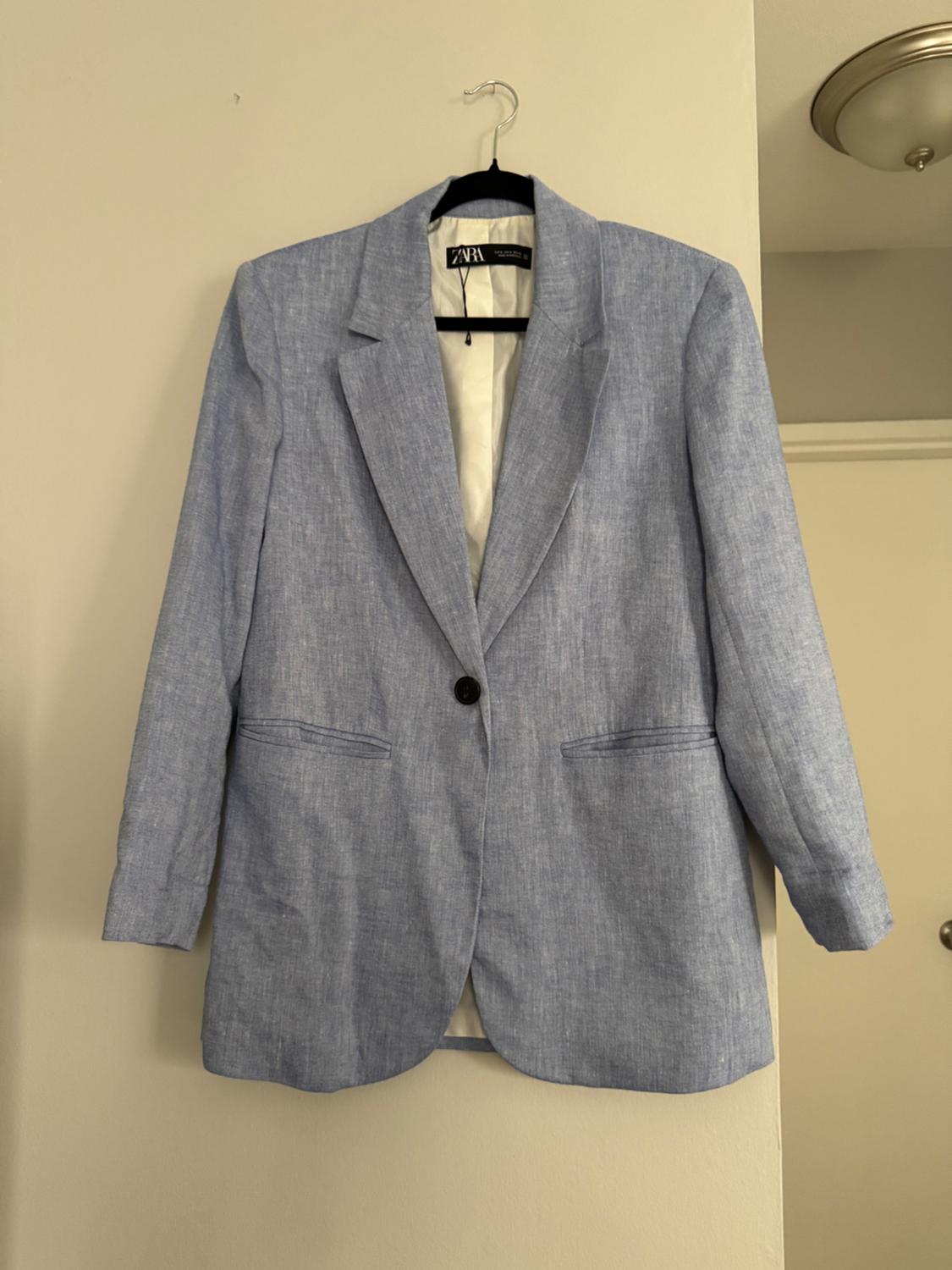 Light blue blazer hanging on a hanger against a beige wall. It has a black button and two front pockets. Label visible on the inside.