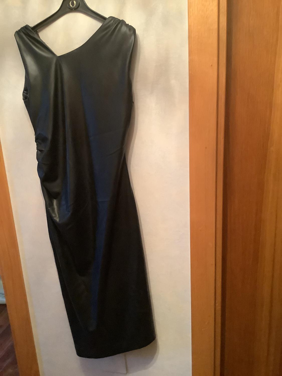 Sleeveless black dress hanging on a hanger, with a draped design on one side, against a light background and wooden frame.