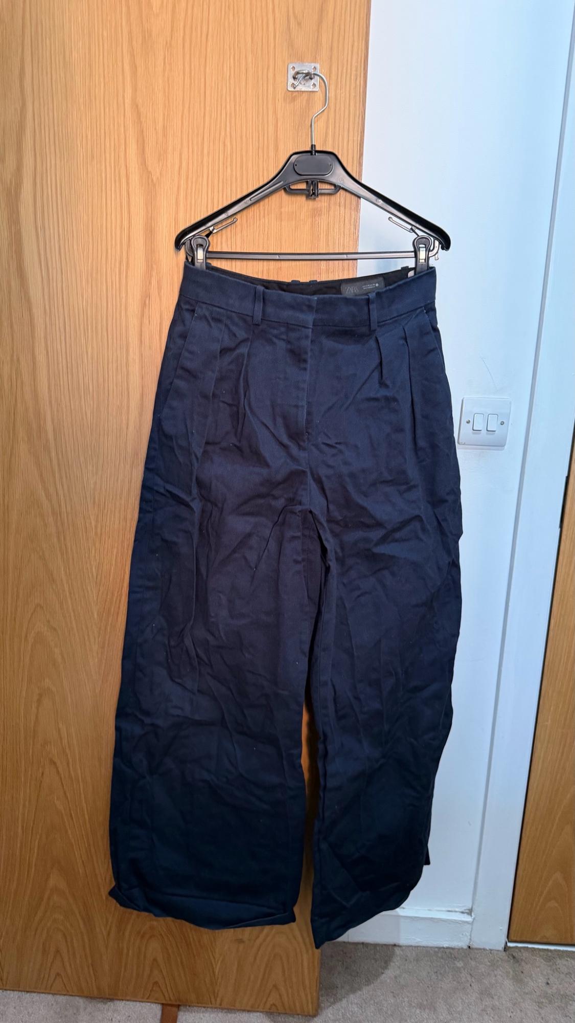 Navy blue wide-leg trousers with pleats, hanging on a coat hanger over a wooden door next to a white wall.