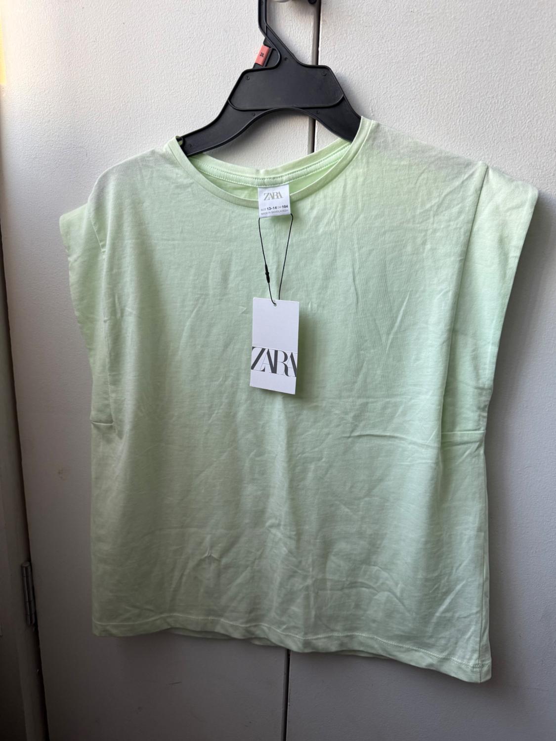 Light green short-sleeved t-shirt hanging on a black hanger against a white background. Tags with the text ZARA and SIZE 13-14 CM 164.