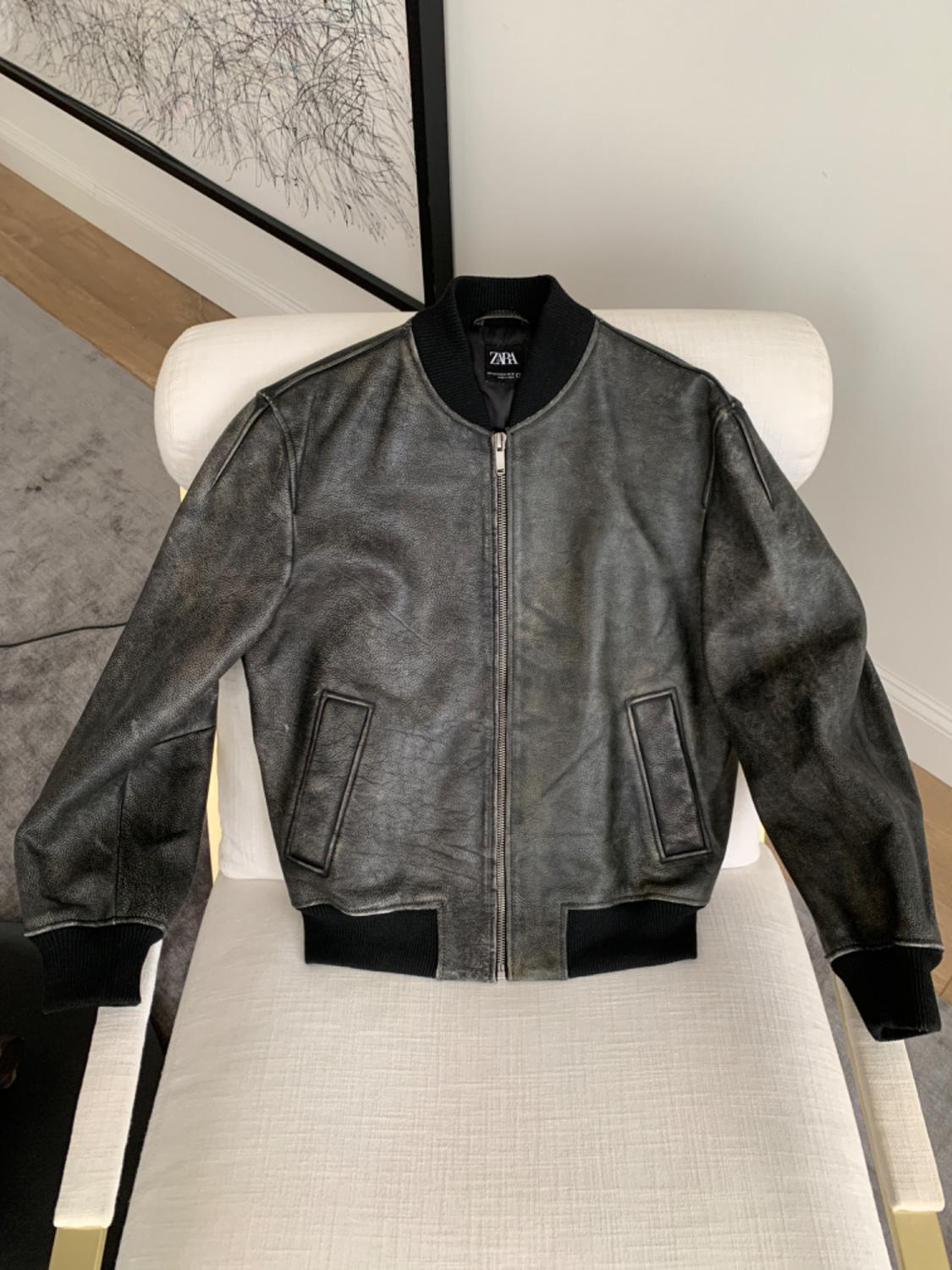 Black leather jacket with a zip and black elasticated cuffs, draped over a white chair. Visible label: ZARA.
