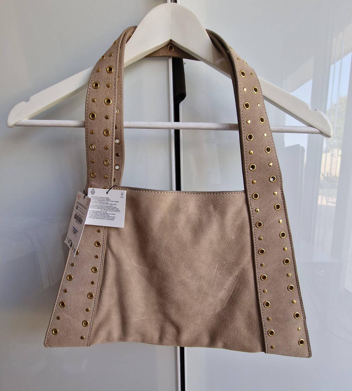 Beige handbag hanging on a white hook. It features wide straps decorated with gold-coloured eyelets and tags.
