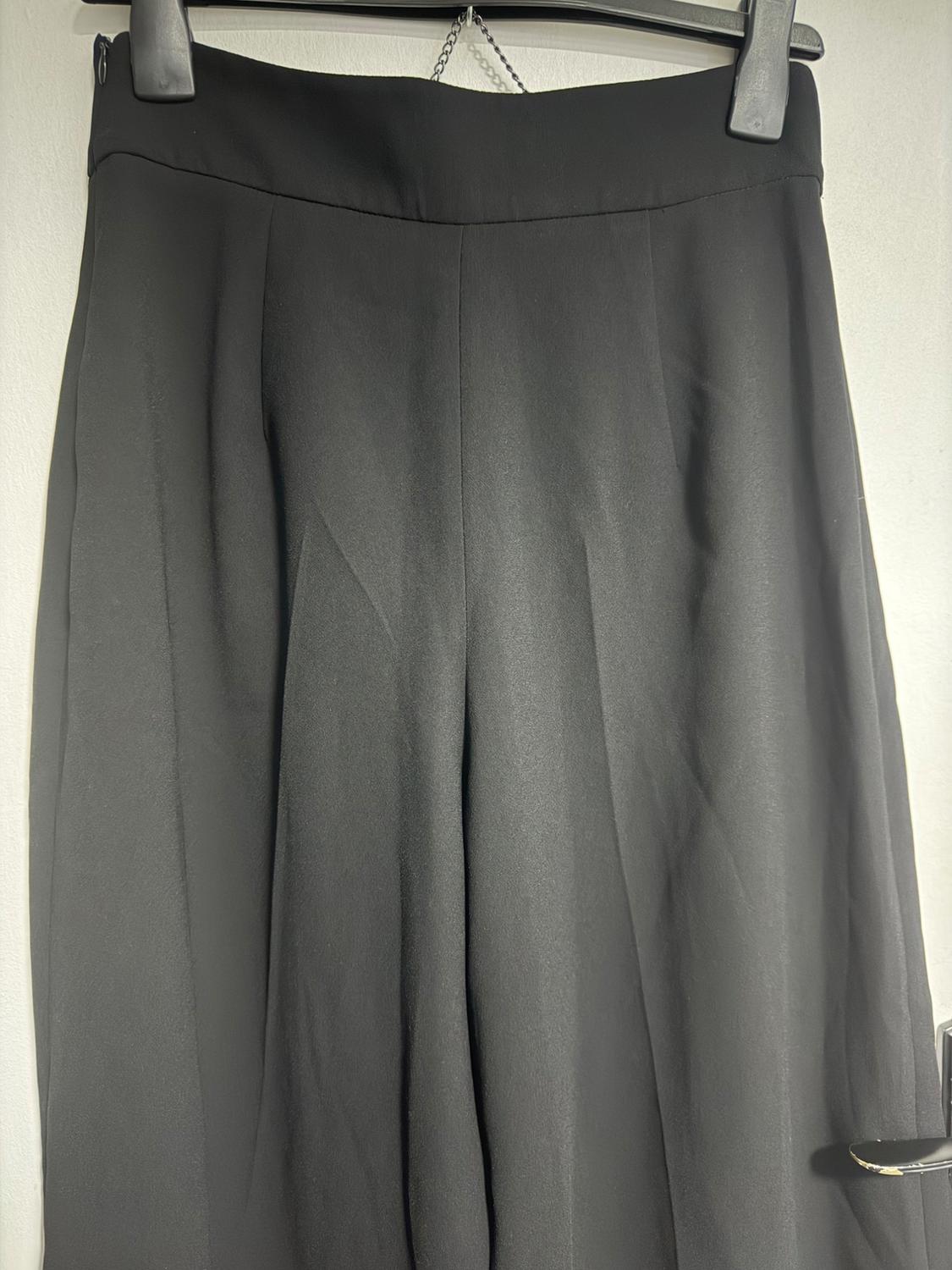 Black trousers hanging on a hanger, with soft pleats and a high waist, against an illuminated white background.