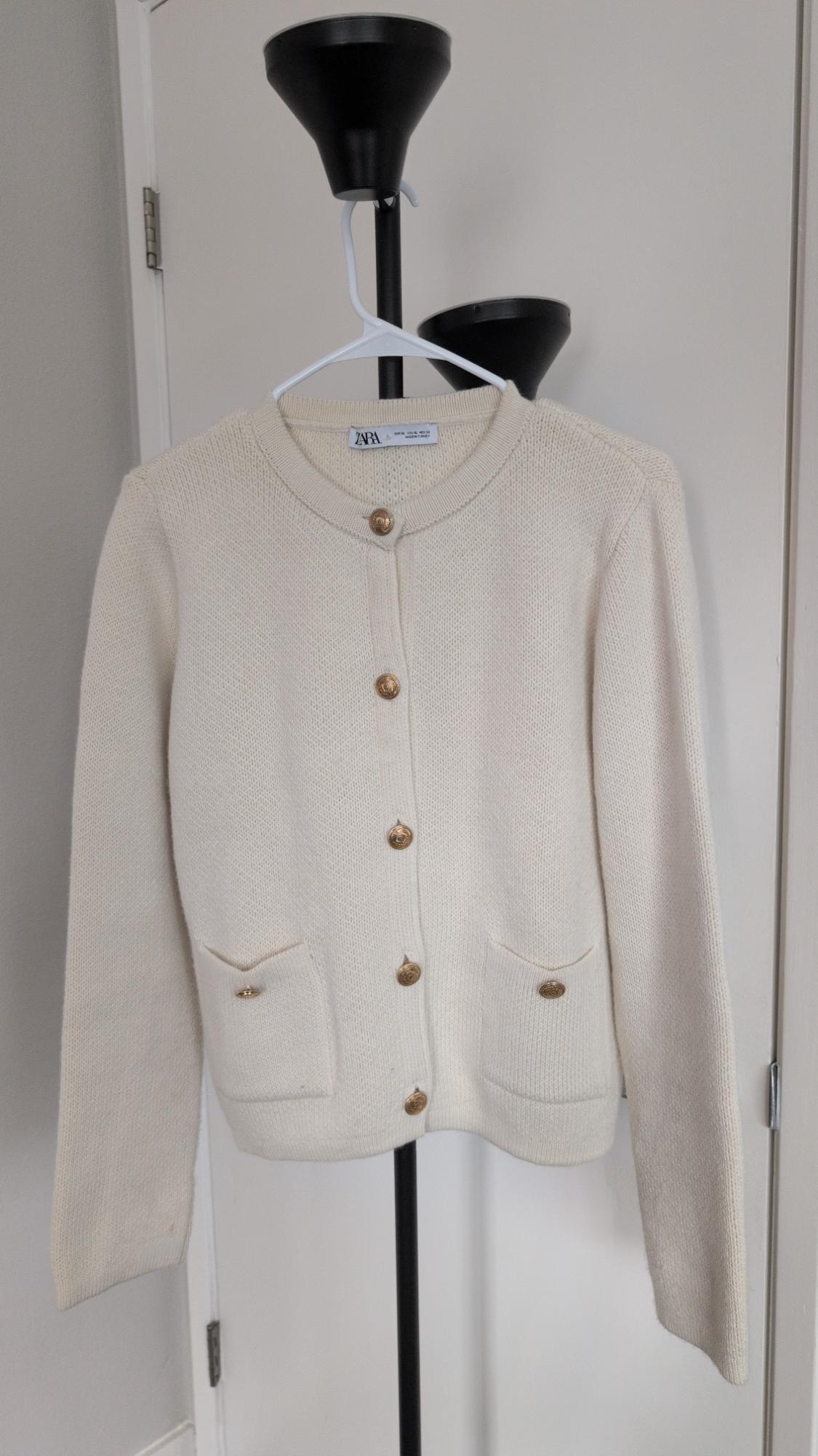 Cream-colored knit cardigan with gold buttons and pockets, hanging on a white hanger. ZARA text visible on the neck label.