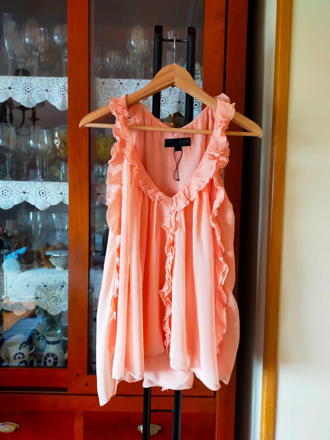 Pink blouse with ruffles hanging on a wooden hanger in front of a glass cabinet with glasses and white lace.