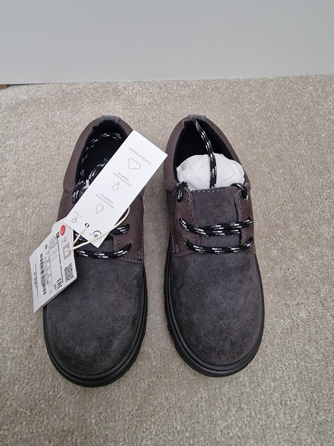 Brown suede shoes with black and white laces, on a beige carpet. Tags hanging from the left shoe.