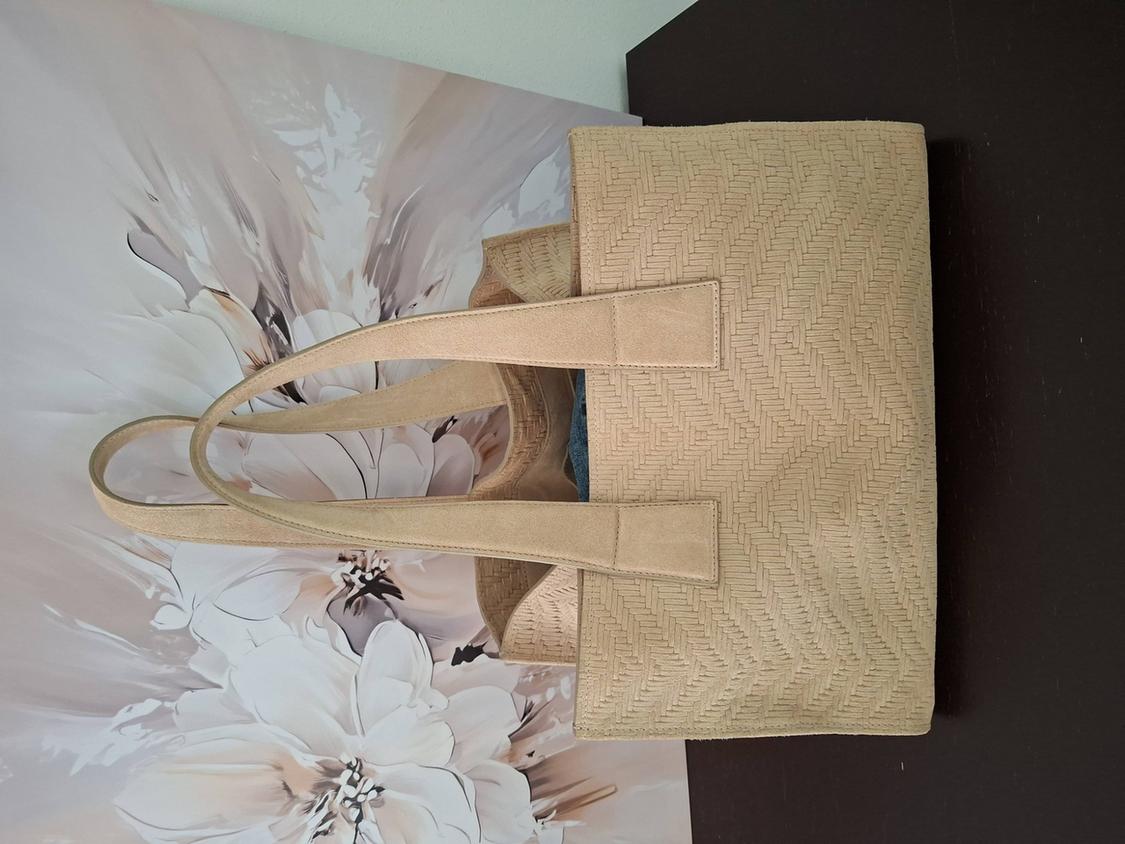 Beige bag with a herringbone texture on a background of white and brown flowers. The bag has long handles and is on a dark surface.