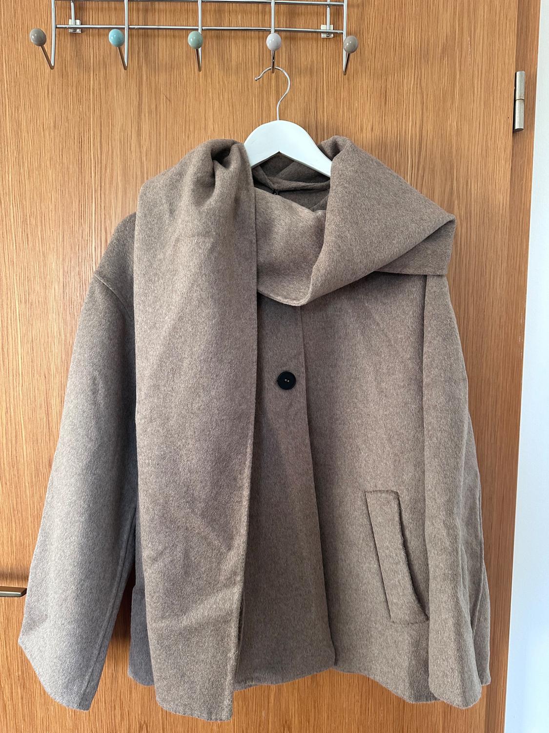 Light brown coat hanging on a white hanger over a wooden door. It has a black button and a side pocket.