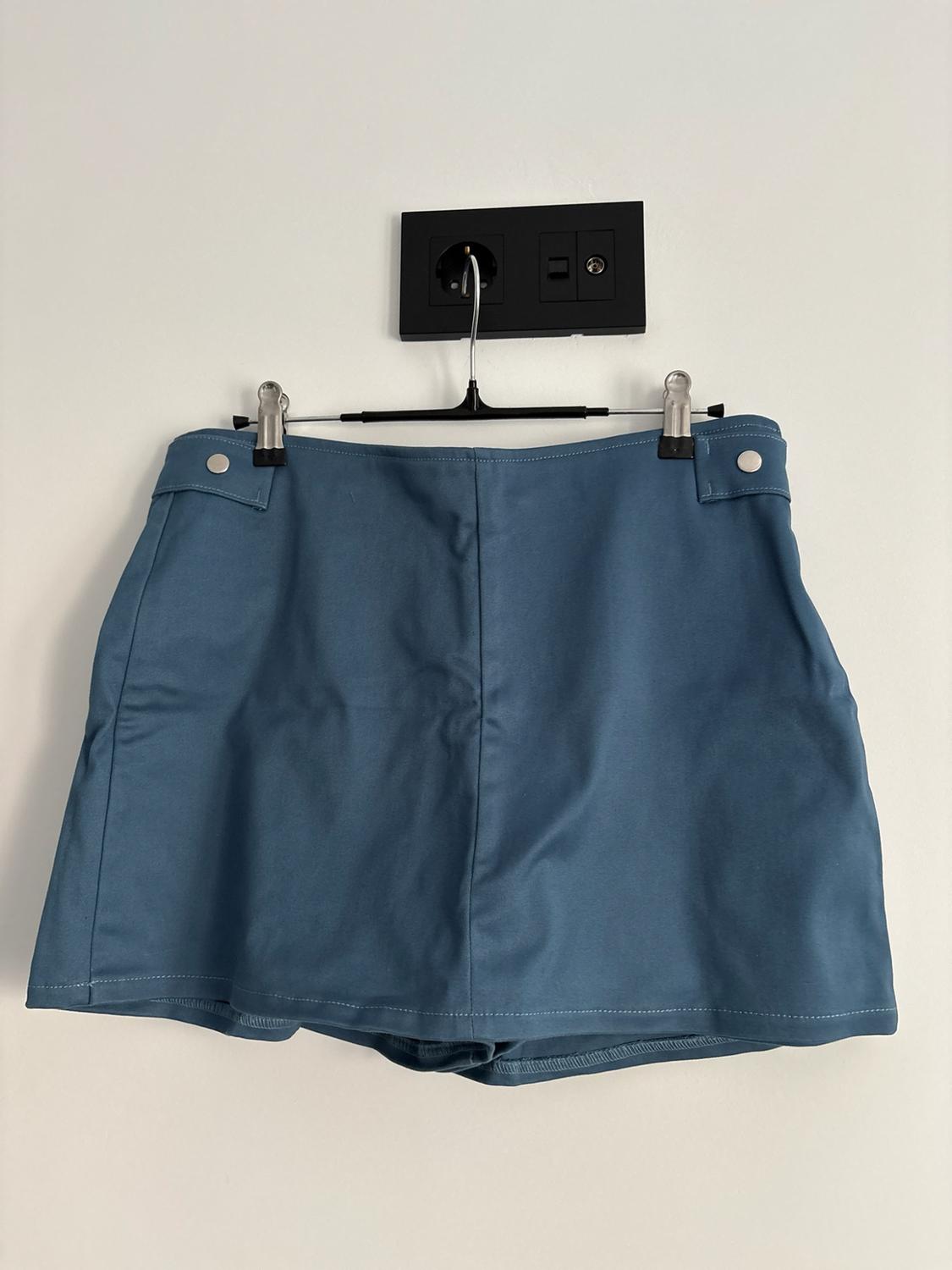 Blue culottes with buttons, hanging from a black hanger attached to a black plug socket on a white wall.