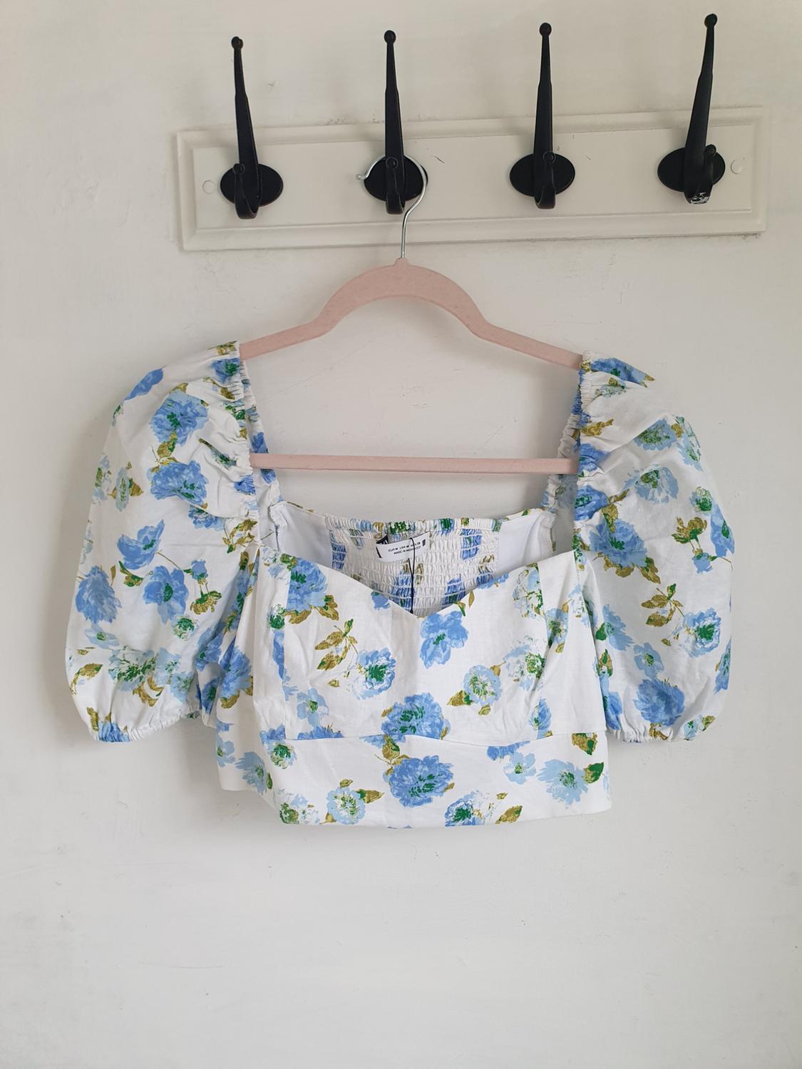 Short-sleeved blouse with blue and green floral print on a white background, hanging on a wooden hanger in front of a white wall.