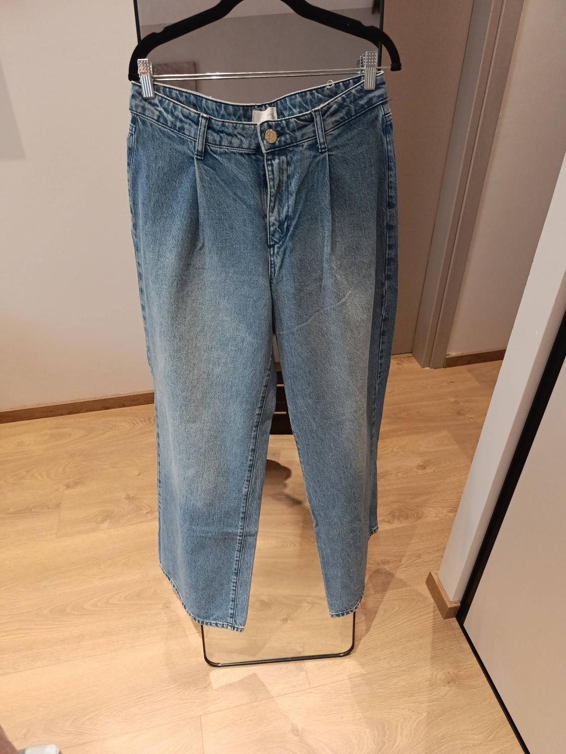 Light blue denim trousers with front pleats, hanging from a black hanger in a room with a wooden floor.