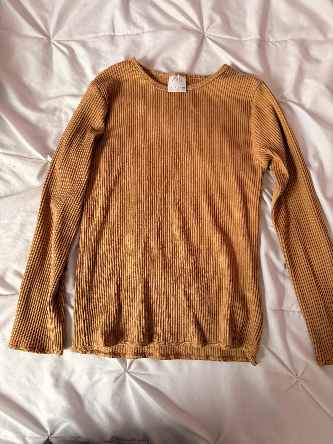 Light brown ribbed knit sweater on a white bedspread. Visible tag on the neck.