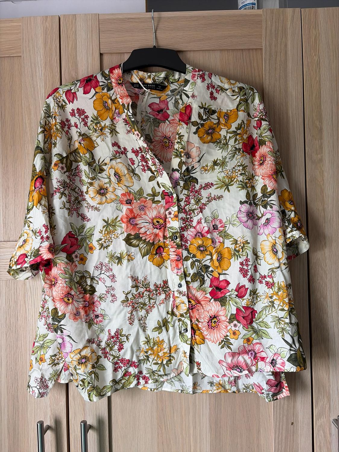 White short-sleeved button-down shirt with a pink and yellow floral print, ZARA BASIC label, hanging in front of a wooden wardrobe.