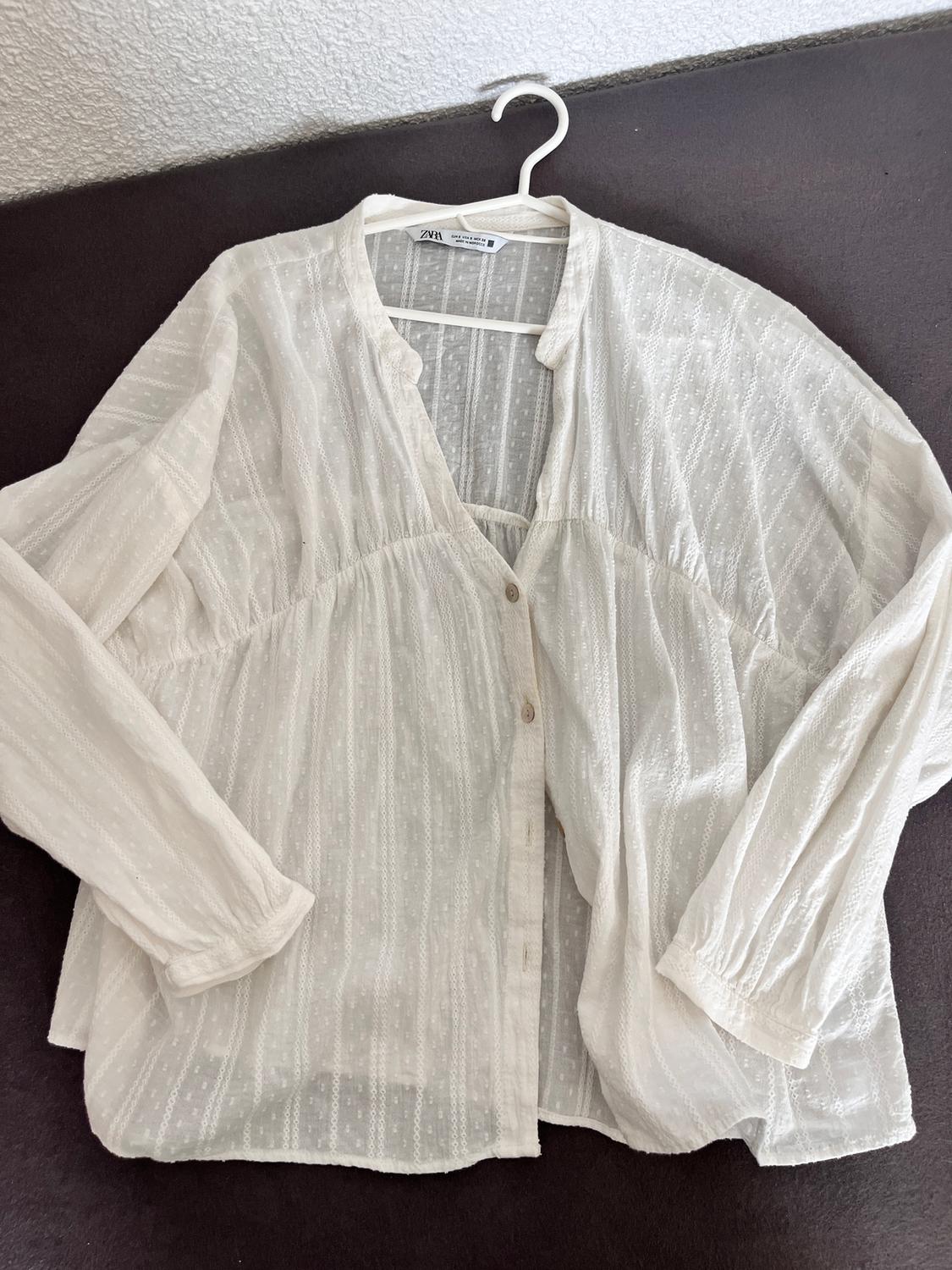 Semi-sheer white blouse with a striped and polka dot pattern, long sleeves, hanging on a white hanger against a dark background.