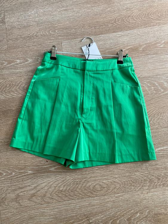 Green shorts hanging on a hanger over a light wooden floor. White tag visible on the hanger.