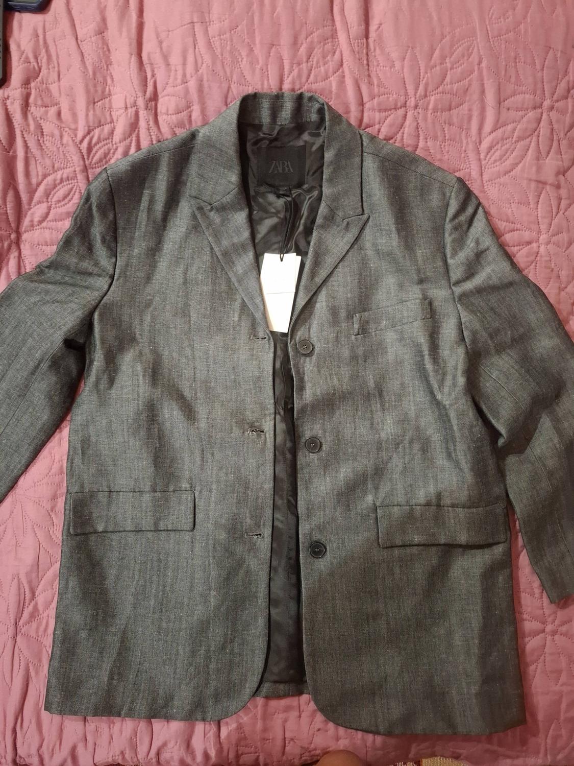 Three-button grey blazer with a hang tag, laid out on a pink quilted bedspread. Inner label bearing the text ZARA.