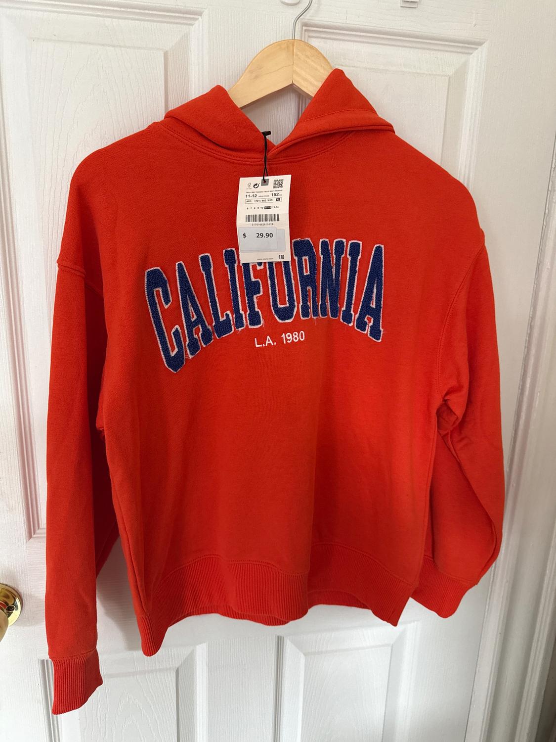 Red hoodie hanging on a white door. Text "CALIFORNIA L.A. 1980" in blue and white. Price tag $29.90.