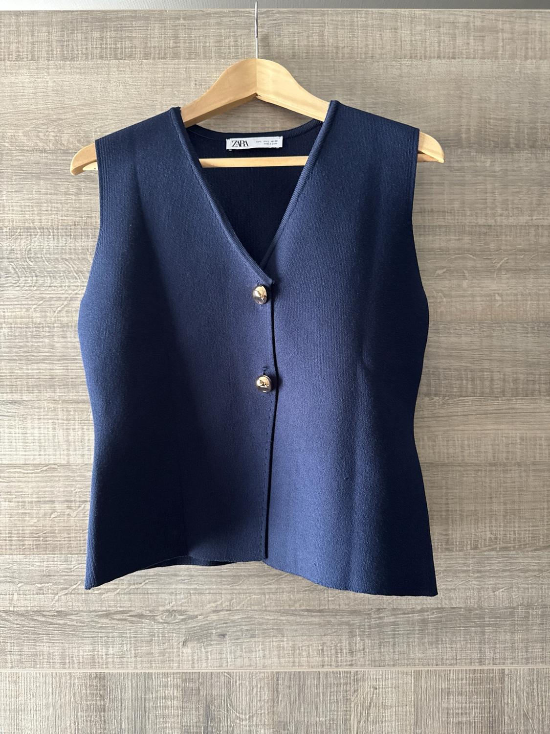 Navy blue knitted V-neck waistcoat with two buttons, hanging on a hanger against a wooden background. The label reads ZARA.