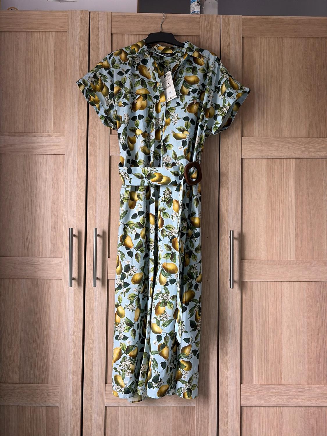 Light blue shirt dress with a lemon print and belt, hanging on a hanger in front of a light wood wardrobe.