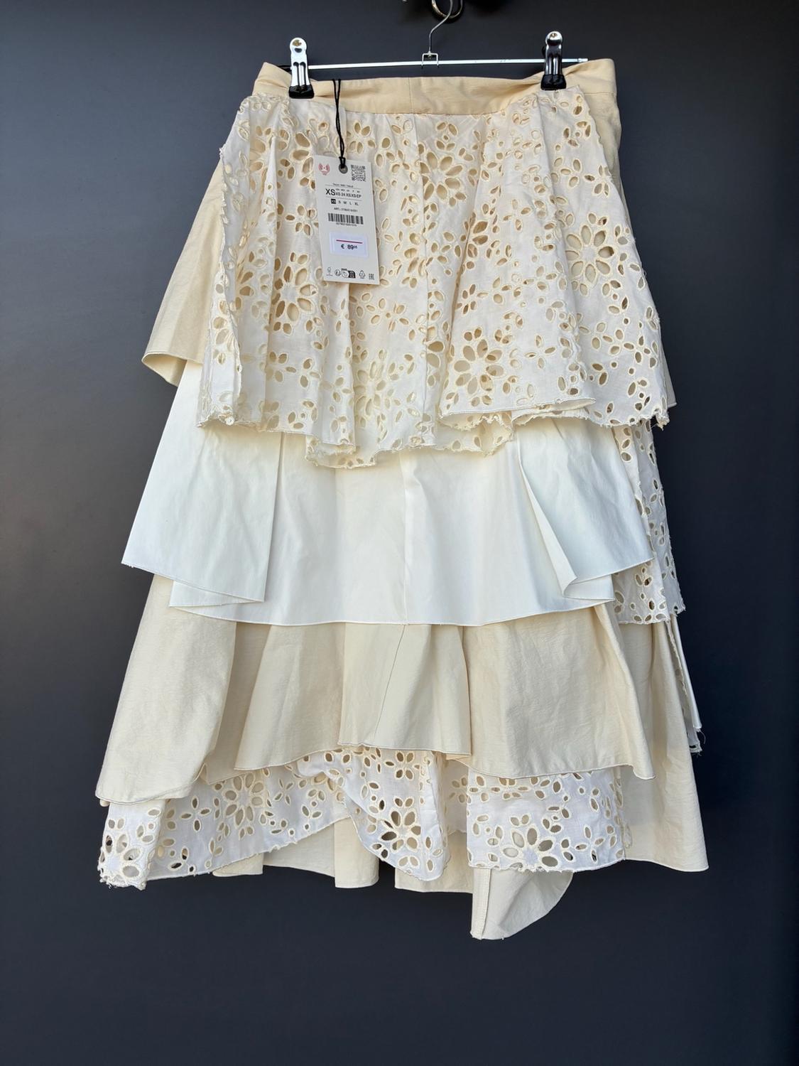 Layered skirt hanging against a dark background. Alternating plain cream and white fabric with openwork details. Visible label displaying XS and €89.
