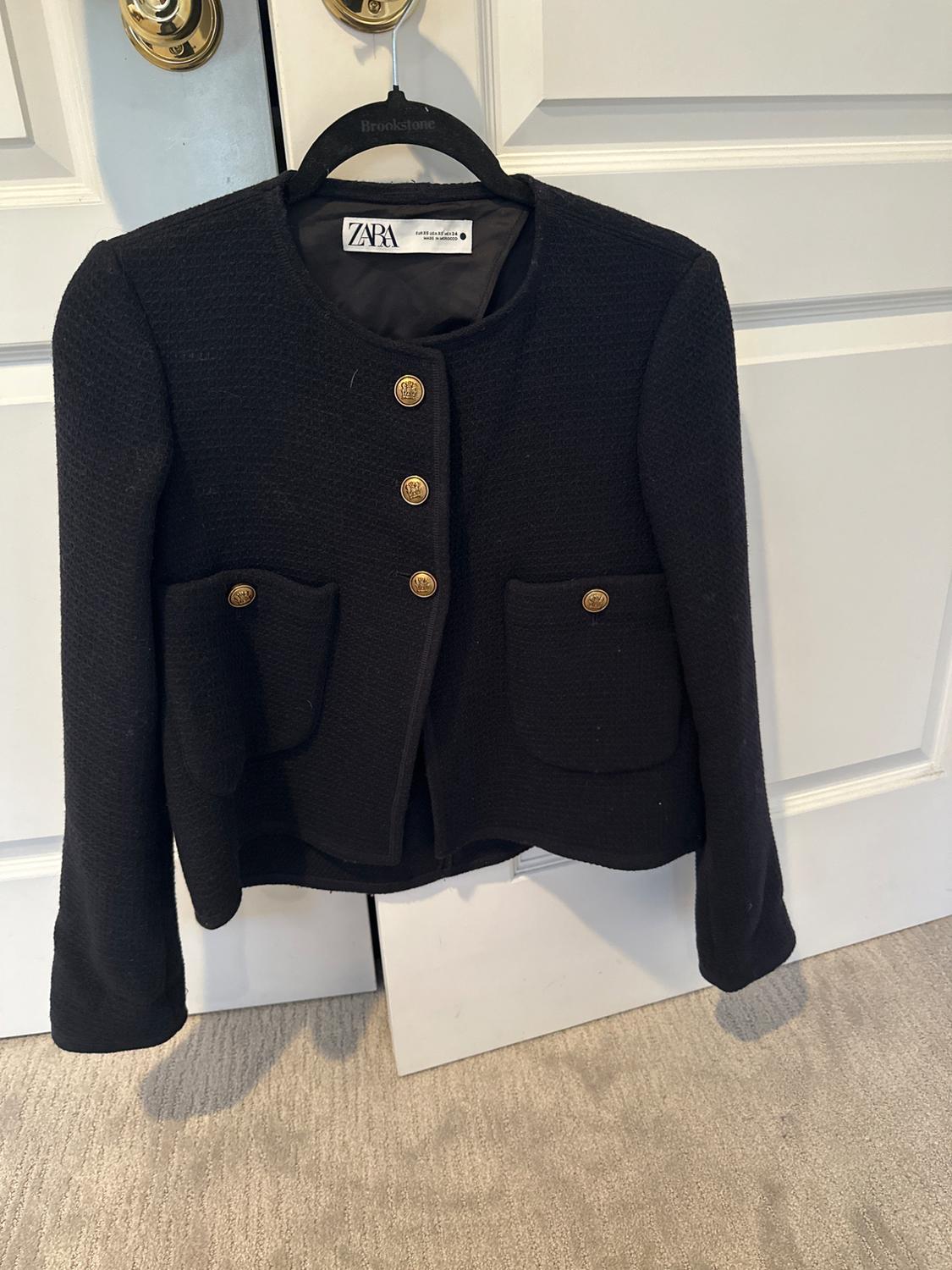 Zara black jacket with gold buttons and front pockets, hanging on a black hanger in front of a white door.