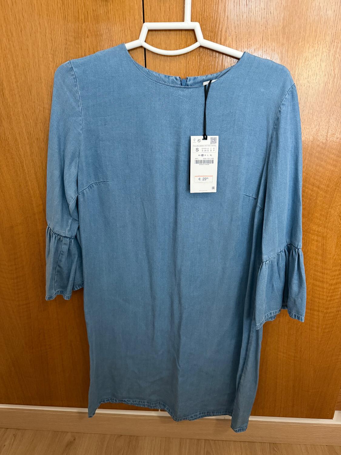 Long-sleeved blue dress hanging on a white hanger, with a visible €29 price tag. Light wooden background.