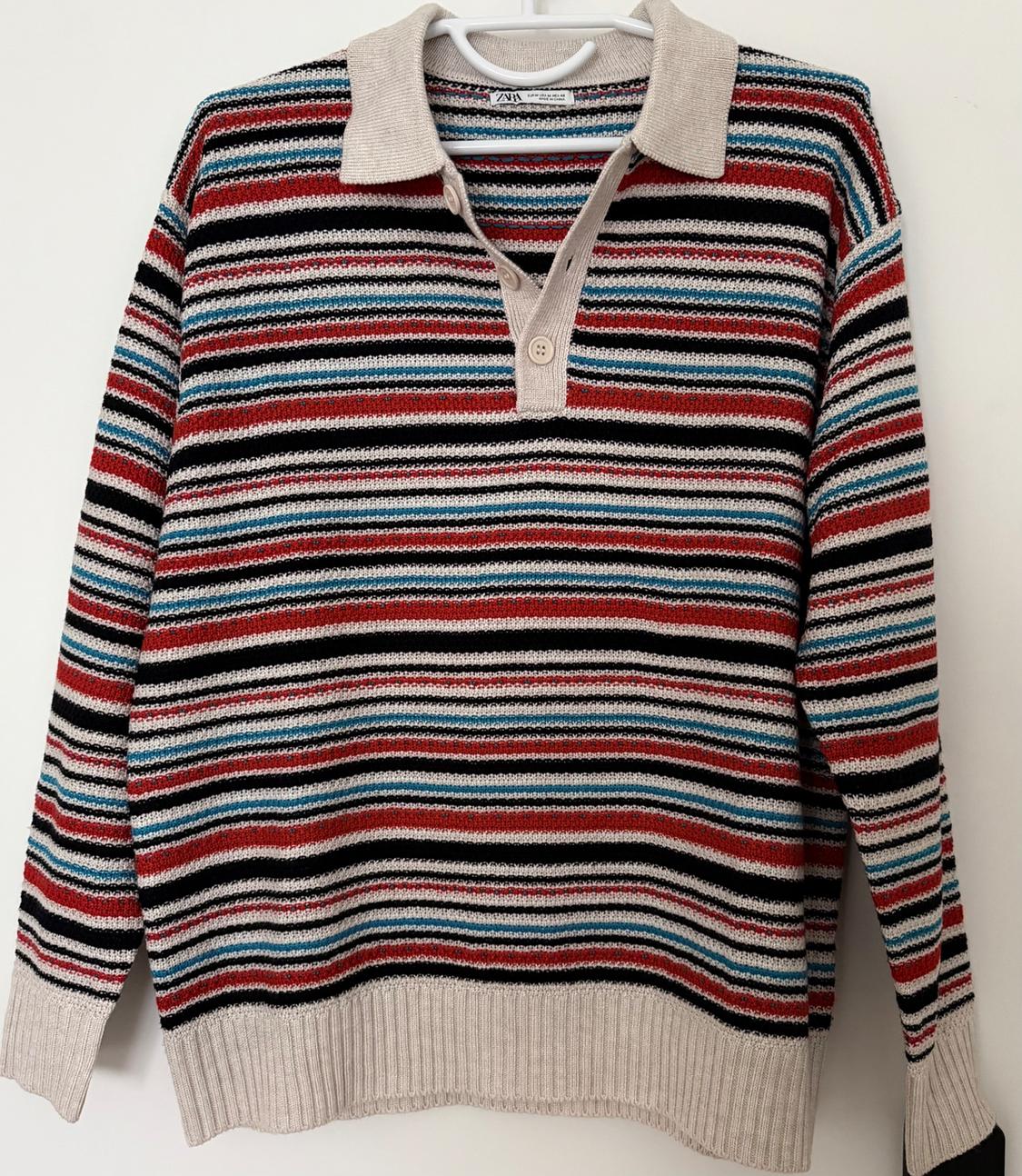 Horizontal striped jumper in red, black, blue, and beige, with a beige collar and cuffs, hanging on a white hanger.