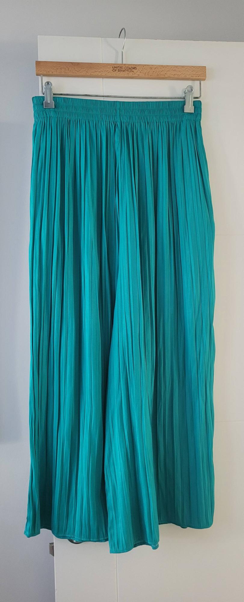 Turquoise pleated trousers hanging on a wooden hanger with the text "United Colors of Benetton" against a white background.
