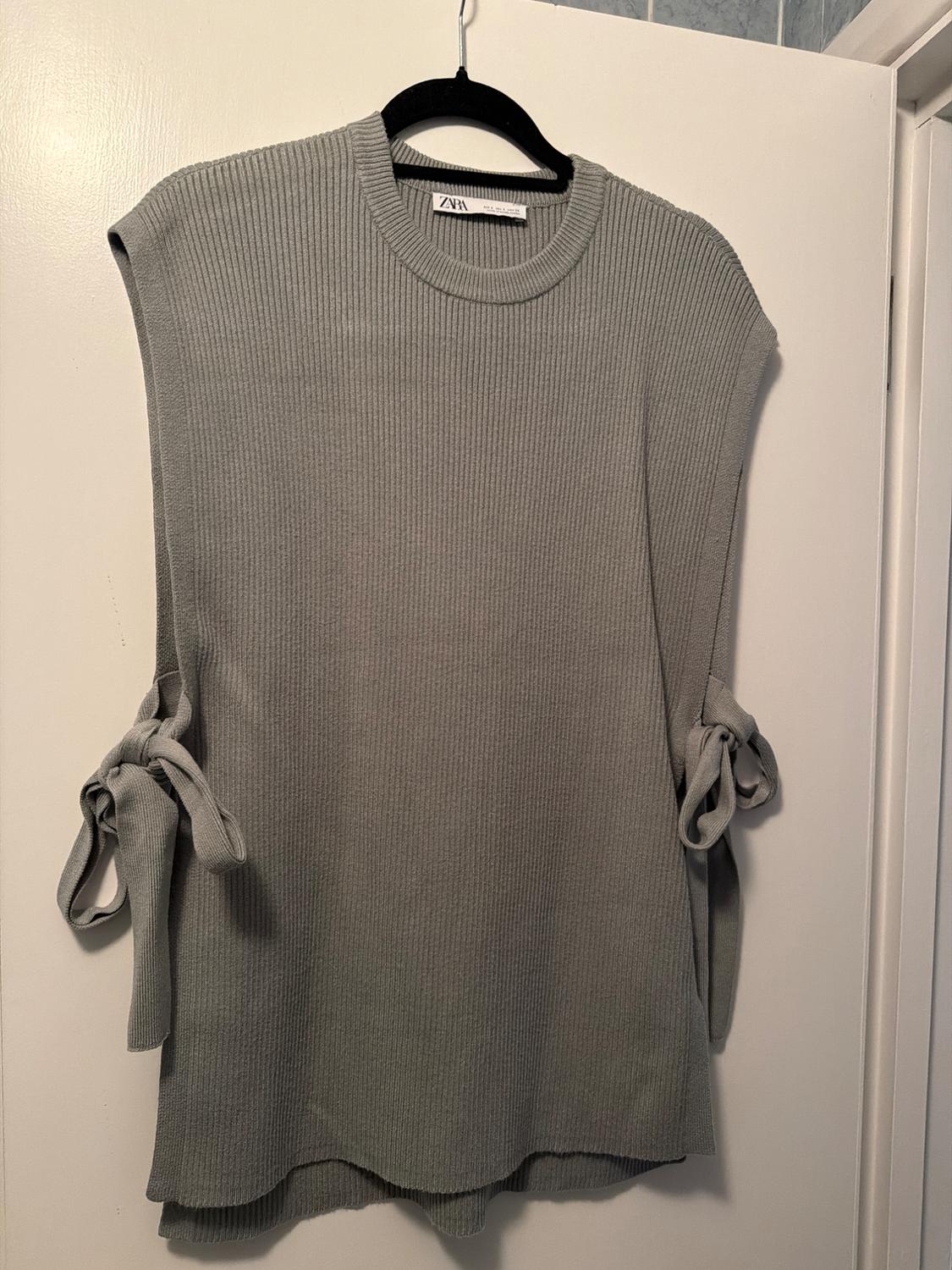 Grey sleeveless jumper with a ribbed texture, ties at the sides, hanging on a black hanger. Visible label: Zara.